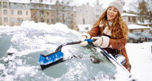 Femme nettoyant la neige du pare-brise d’une voiture avec une brosse pendant l’hiver.